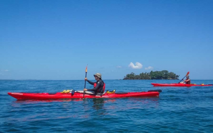 Two red kayaks are paddled by outward bound students on very blue water, with a blue sky above. There is a small tree-covered island in the background.